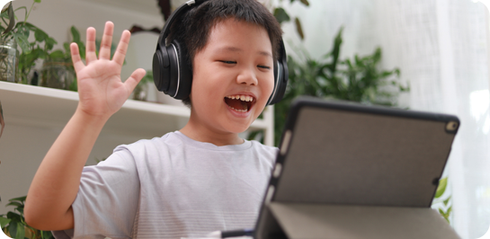 image of young boy on smart device with headphones on. The young boy is smiling at the screen and holding up his hand.